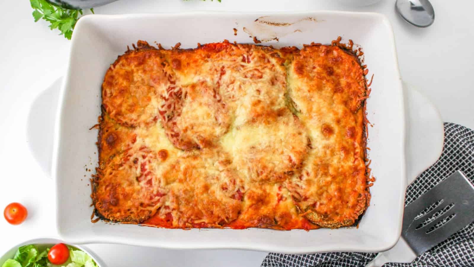 Baked eggplant parmesan in a white casserole dish, topped with melted cheese and tomato sauce.