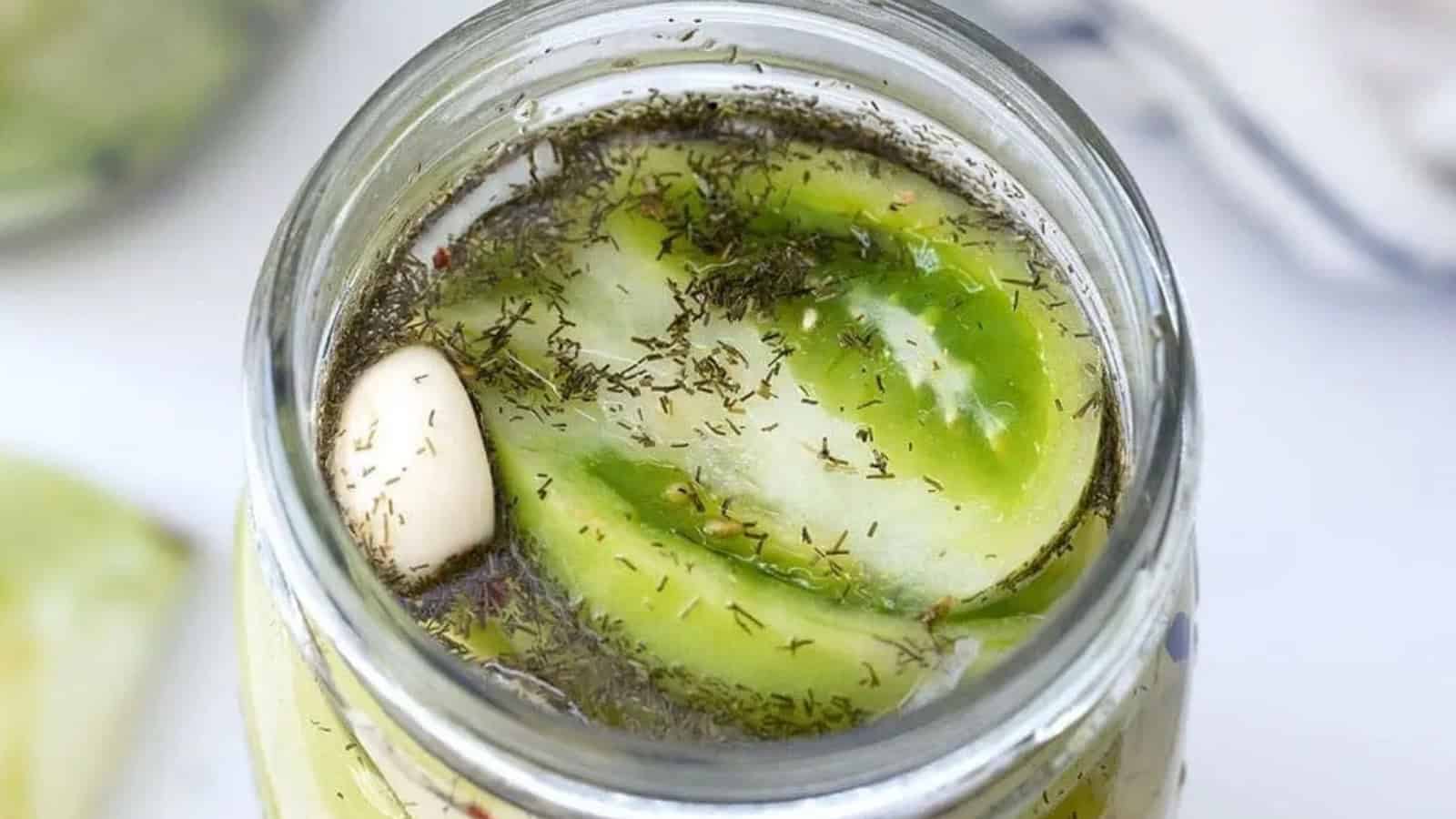 Sliced green tomatoes, garlic, and herbs in brine inside a glass jar, viewed from above.