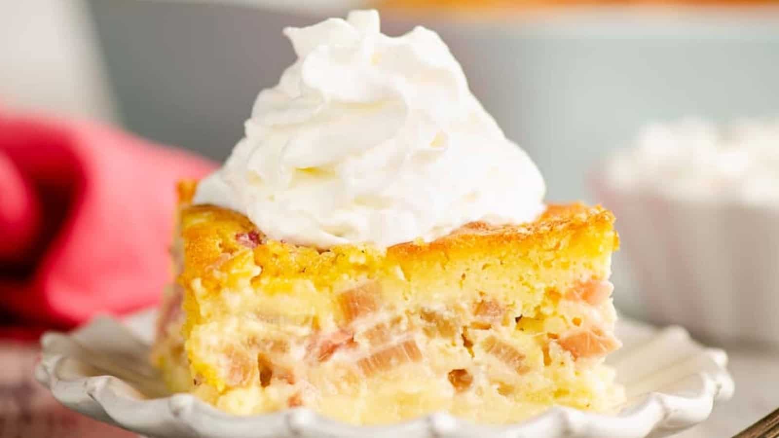 A slice of rhubarb custard cake topped with a swirl of whipped cream on a white plate.