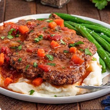 Salisbury steak with gravy and vegetables on mashed potatoes, served with green beans on a white plate.
