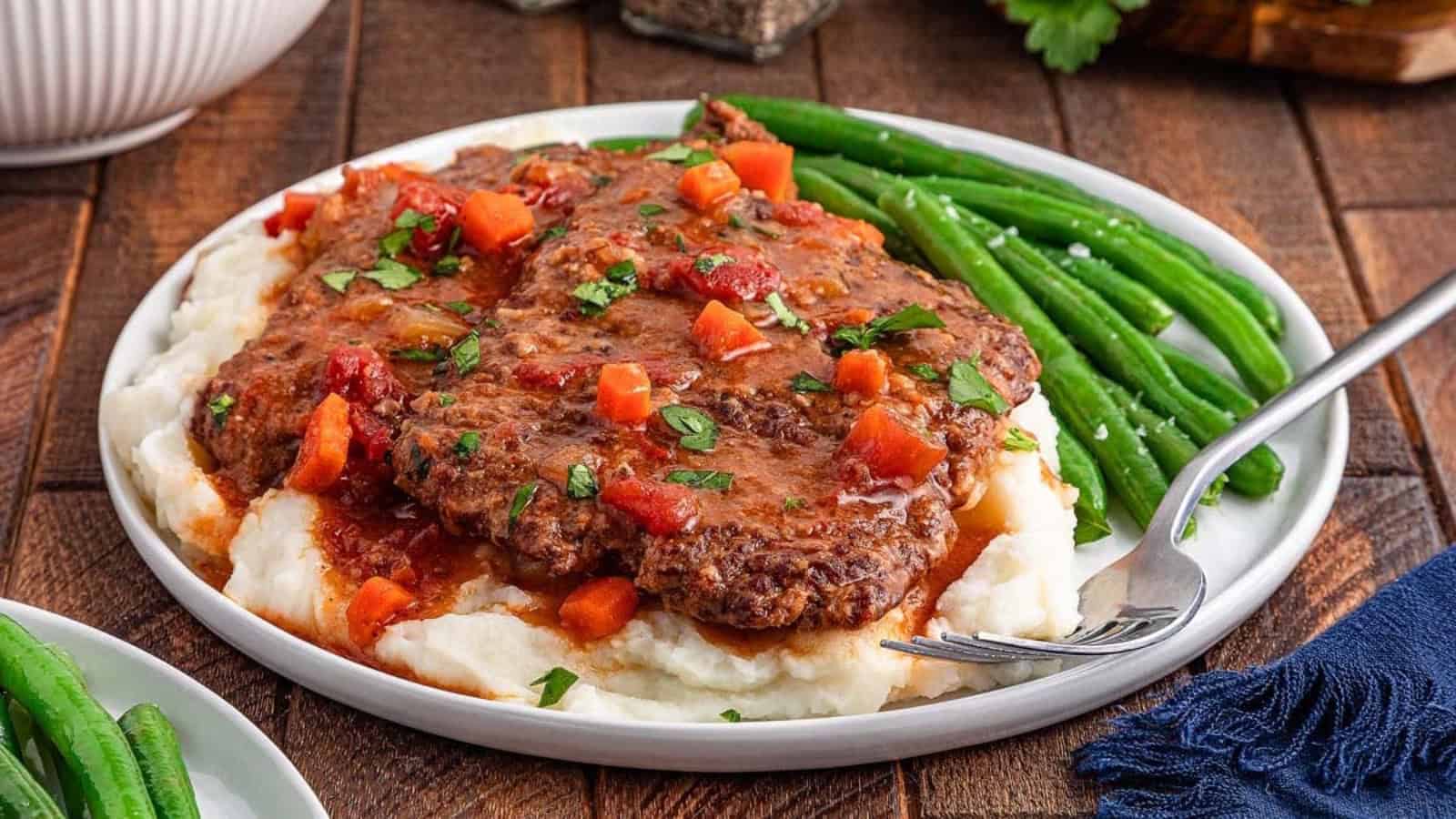 Beef steaks in tomato gravy served over mashed potatoes with green beans on a plate.