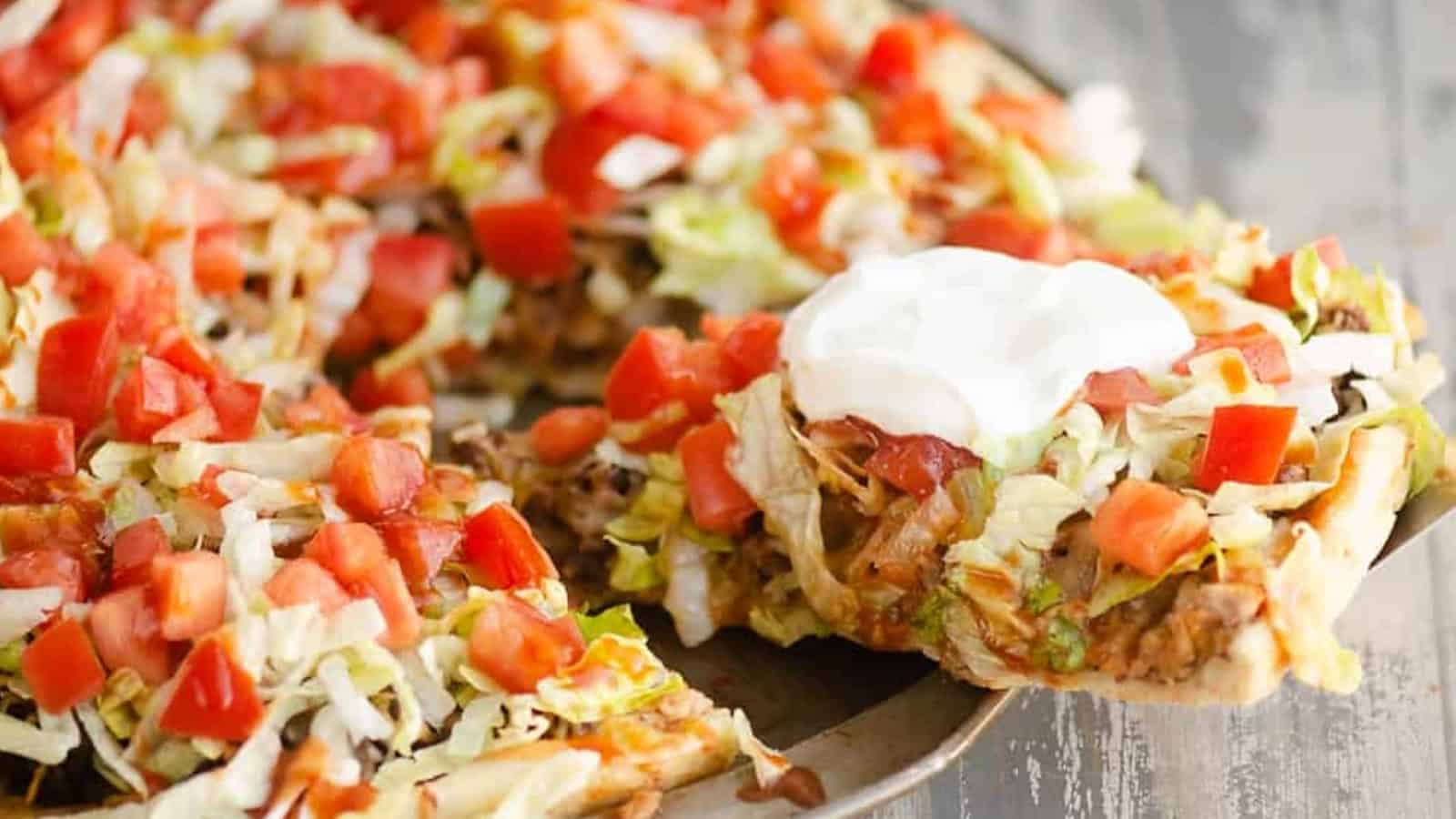 A slice of taco pizza topped with lettuce, tomatoes, and sour cream is being served from a pizza pan.
