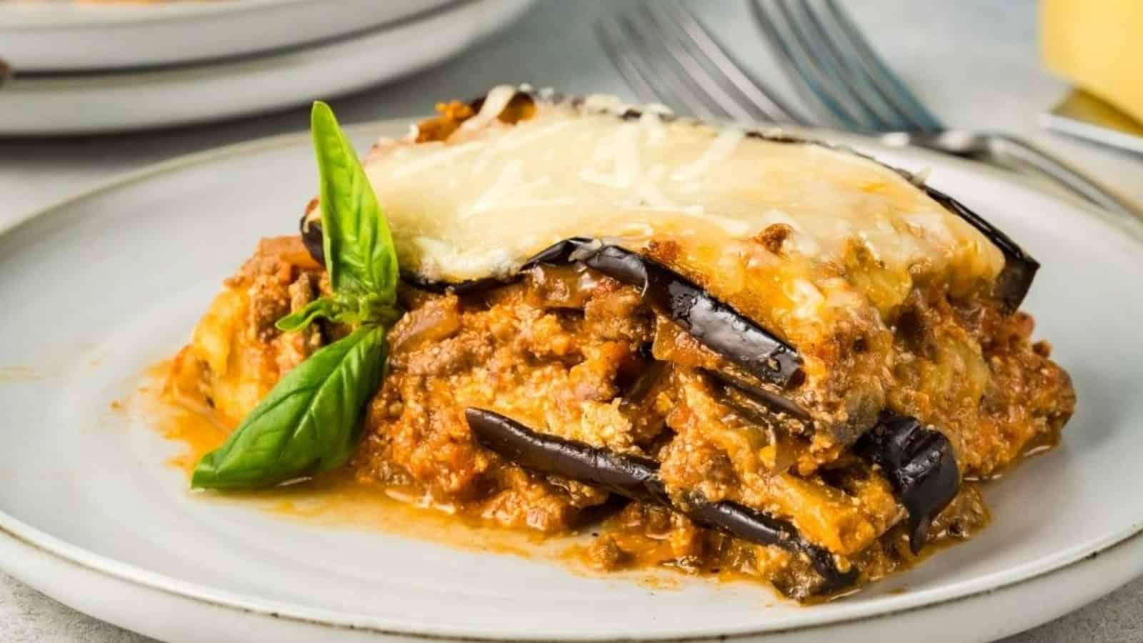 A slice of eggplant lasagna with melted cheese, meat sauce, and a basil garnish on a white plate.