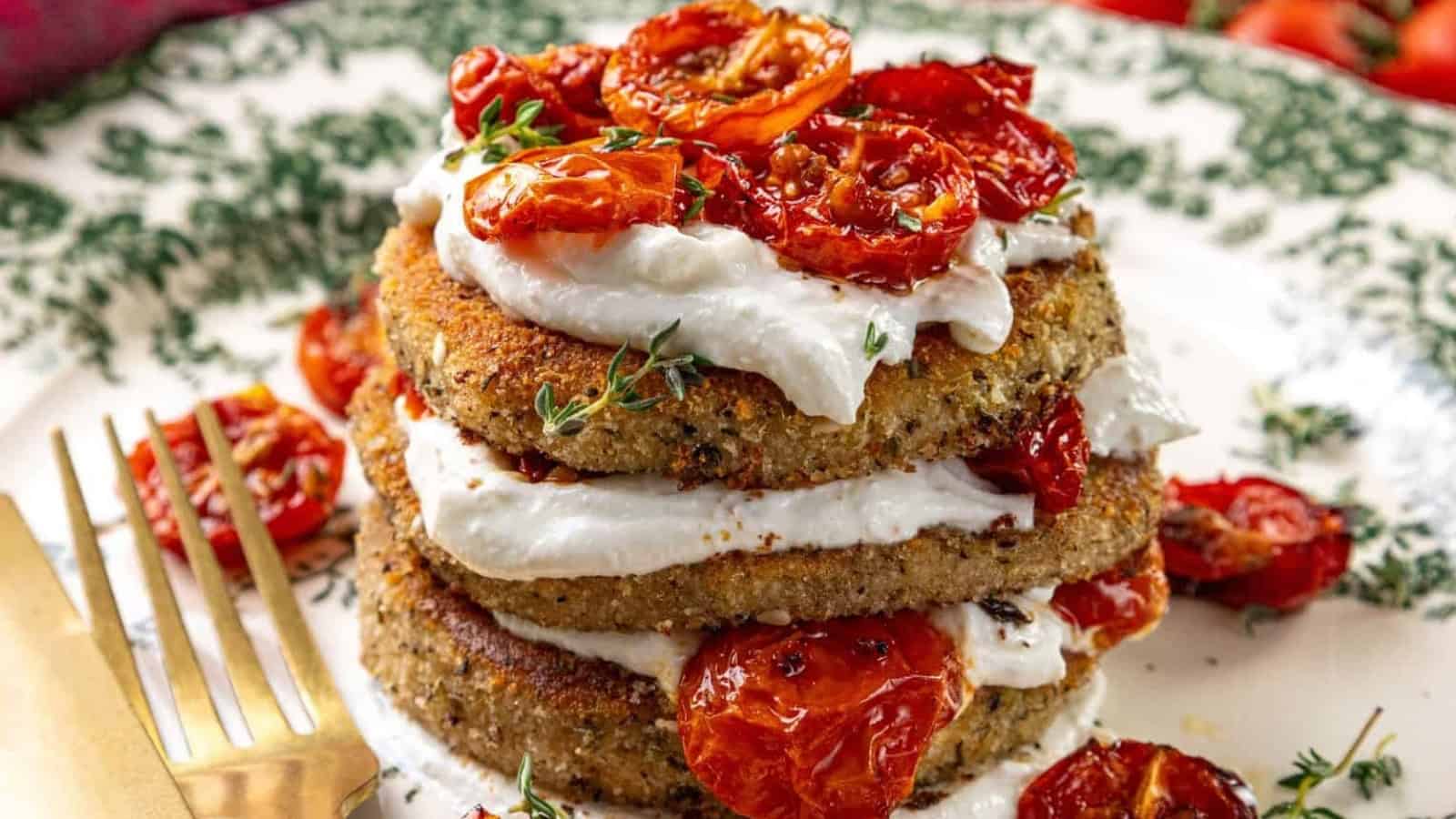 Stack of breaded eggplant slices layered with whipped feta and roasted tomatoes on a decorative plate, with a fork and knife on the side.