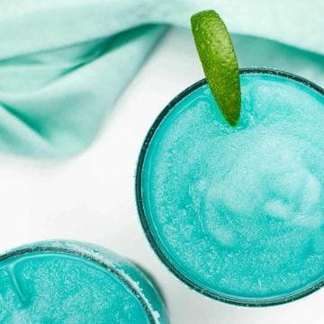 Two bright blue frozen cocktails, one garnished with a lime wedge, next to a light blue cloth.