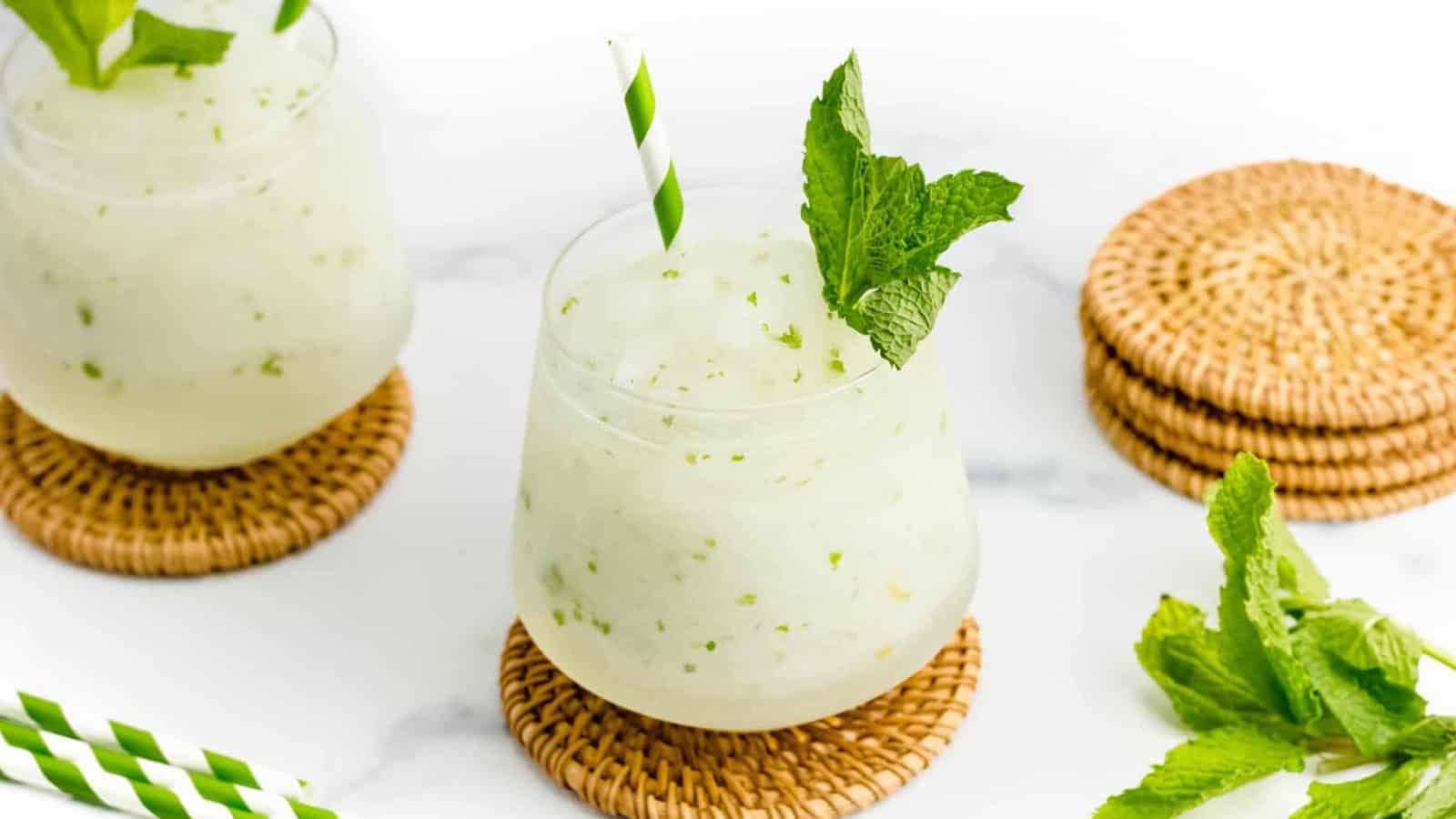 A frosty mint drink with a green-striped straw, garnished with mint, sits on a woven coaster.