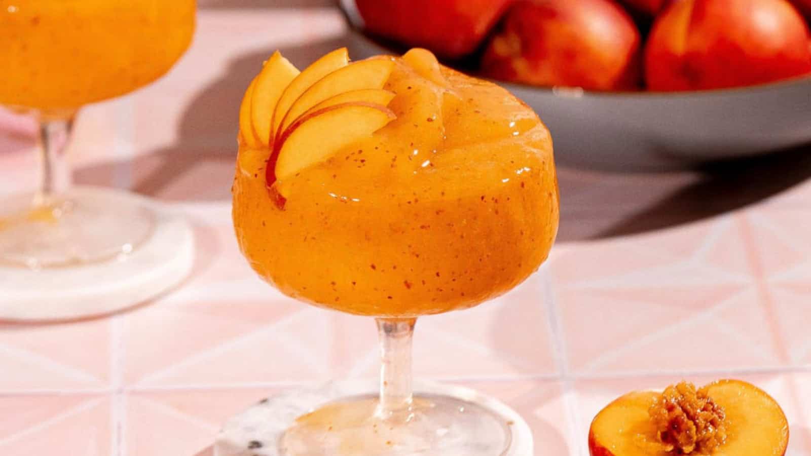 A peach frozen cocktail in a glass, garnished with peach slices, with fresh peaches in the background.