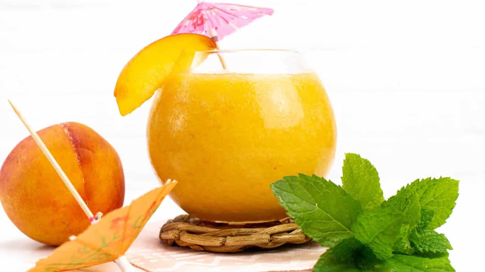 Peach smoothie in a round glass with a peach slice, mint leaves, and cocktail umbrellas for decoration.