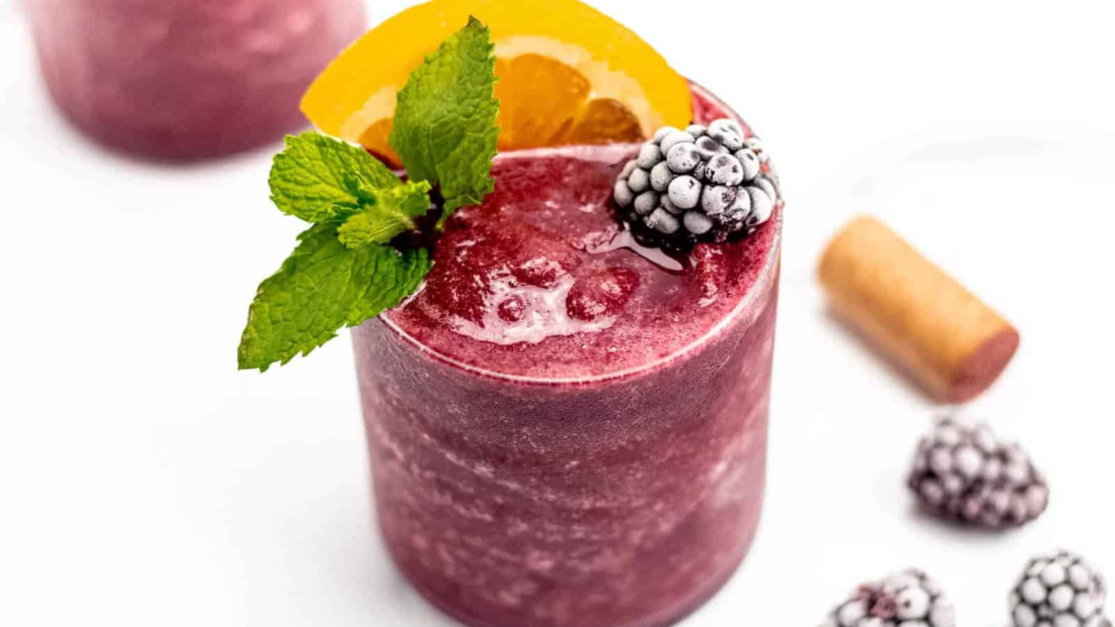 A purple smoothie garnished with a mint sprig, orange slice, and blackberries, with ice and a cork nearby.