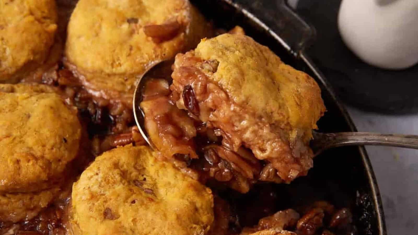 A spoon lifts a serving of pecan and apple cobbler with biscuits from a skillet.