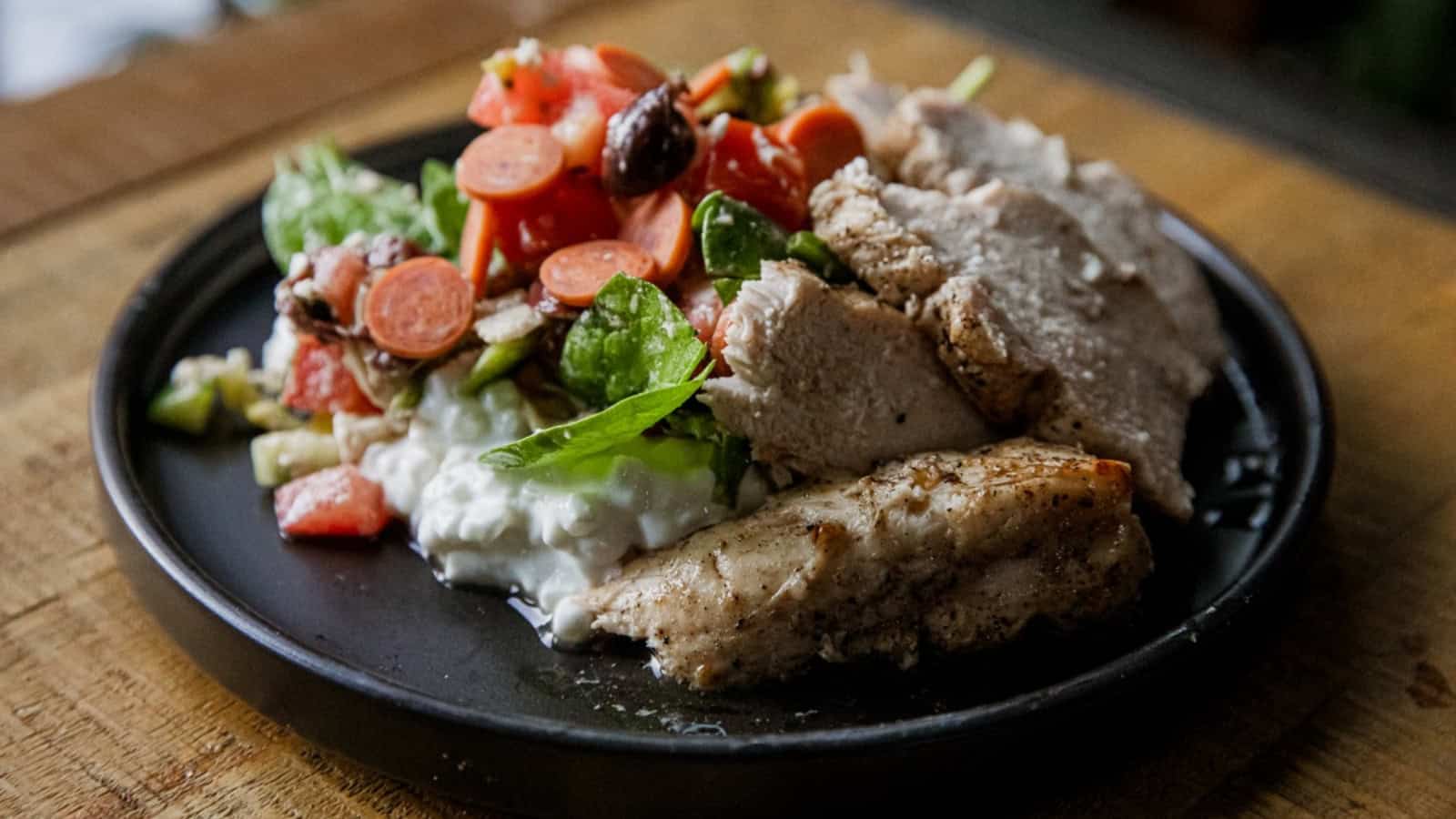A black plate with grilled chicken, cottage cheese, and a colorful salad with tomatoes, carrots, and spinach.