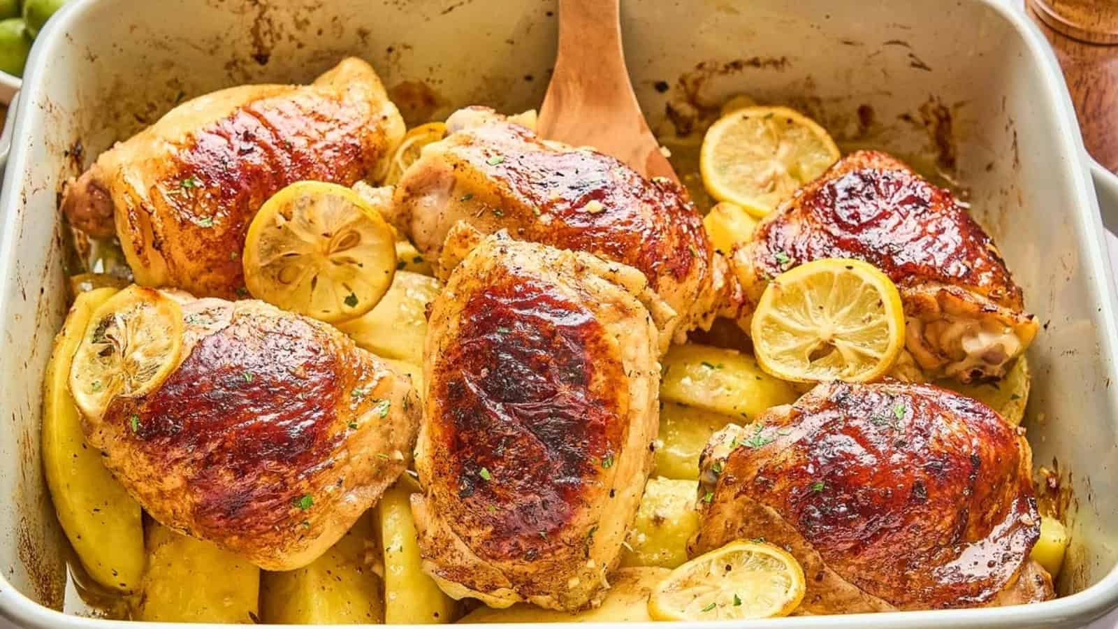 Roasted chicken thighs with golden brown skin, sliced lemons, and potato wedges in a baking dish.