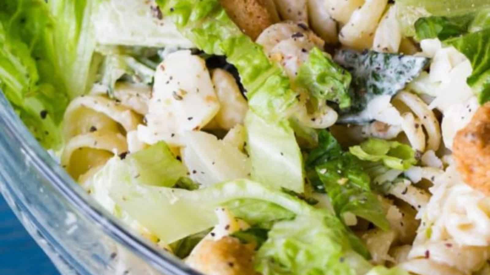 Close-up of a Caesar salad with lettuce, pasta, chicken, croutons, and creamy dressing in a glass bowl.