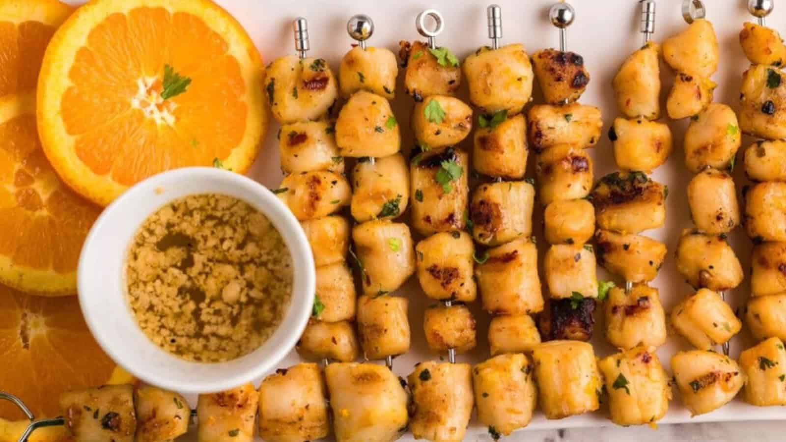 Grilled chicken skewers with parsley, orange slices, and a bowl of crushed peanuts on a white plate.