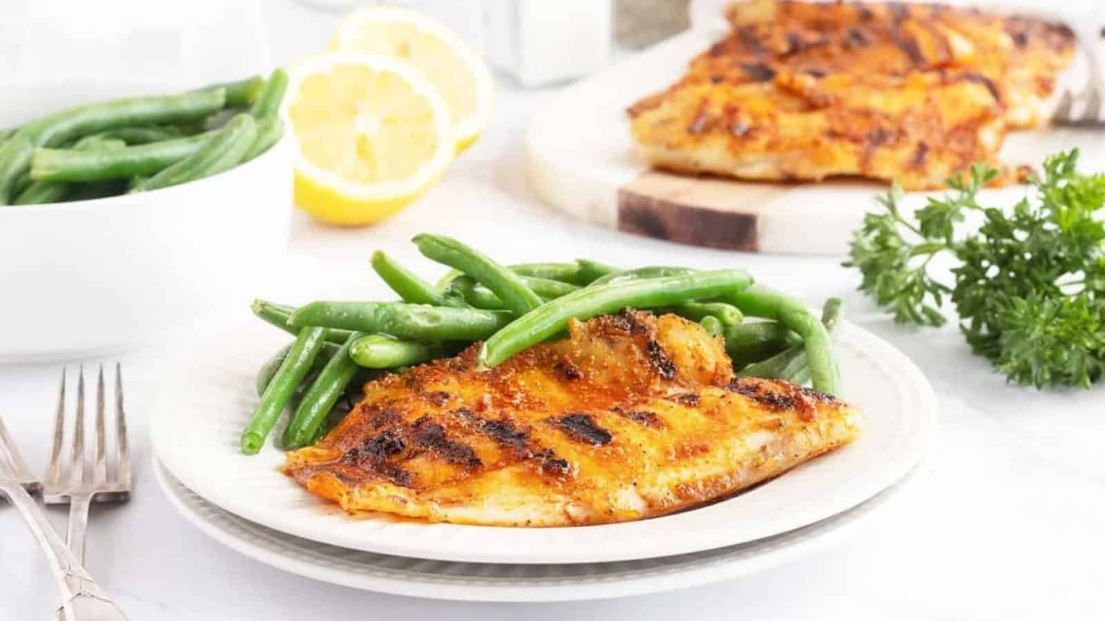 Grilled fish fillet and green beans on a white plate, with lemon and herbs in the background.