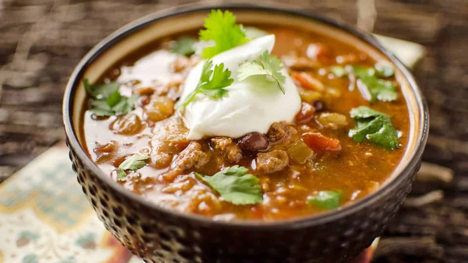 Bowl of chili topped with sour cream and cilantro, with beans, vegetables, and ground meat visible.