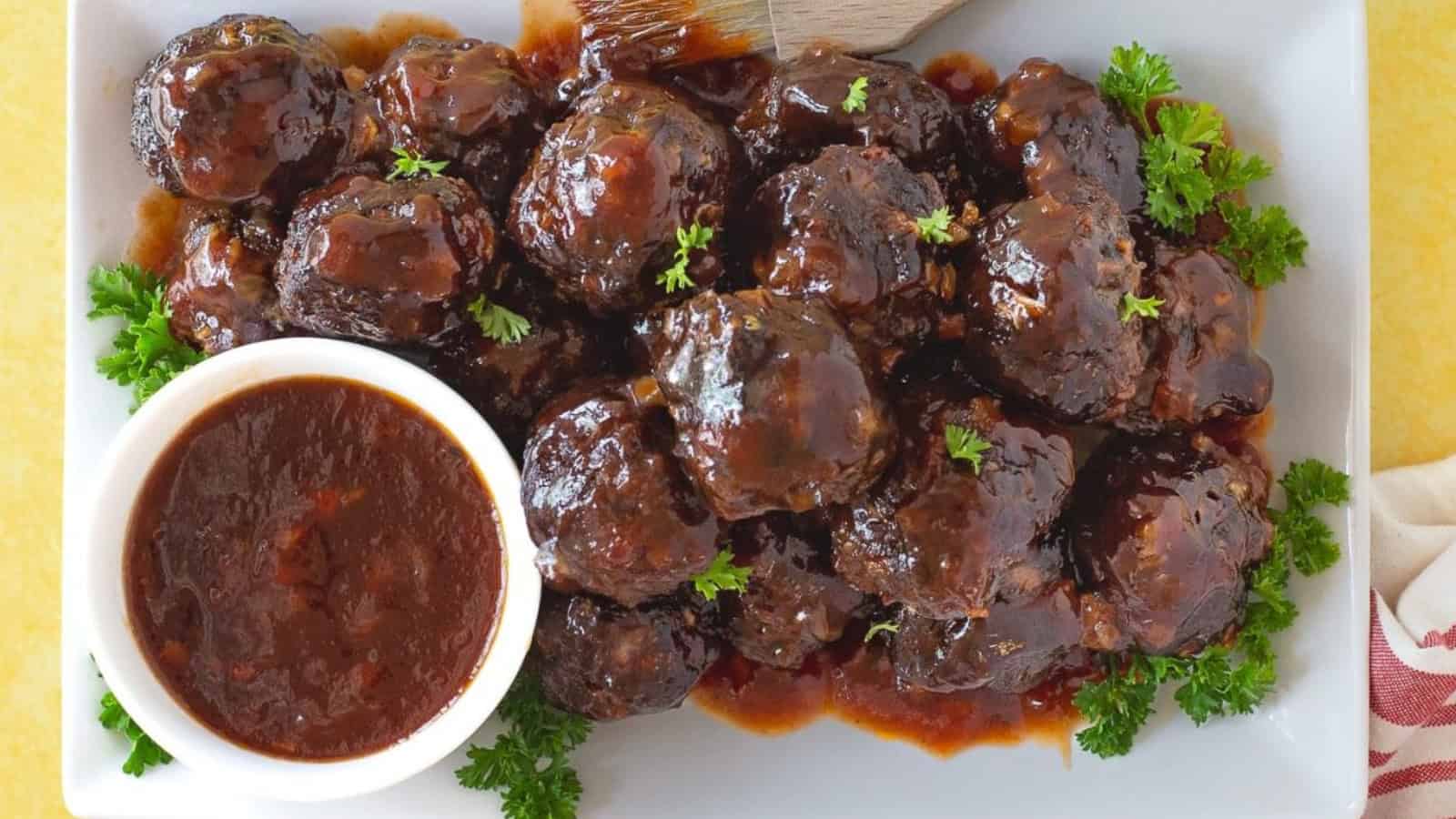Meatballs covered in sauce on a white plate, garnished with parsley, with a bowl of extra sauce on the side.