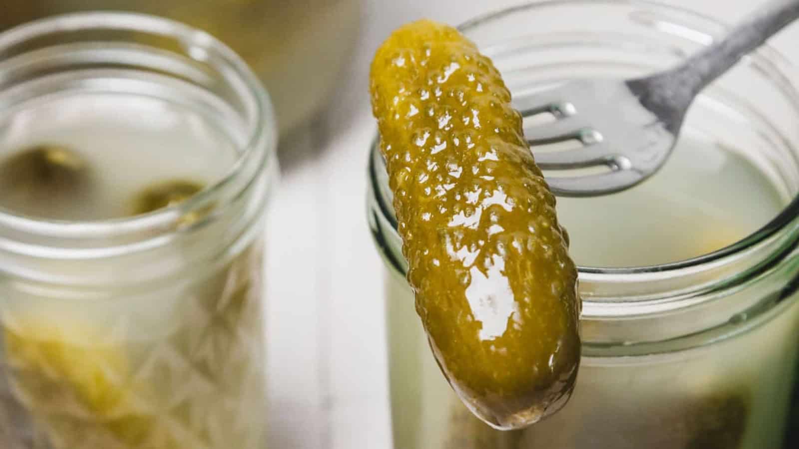 A fork holding a pickle above an open jar of pickles, with another jar in the background.