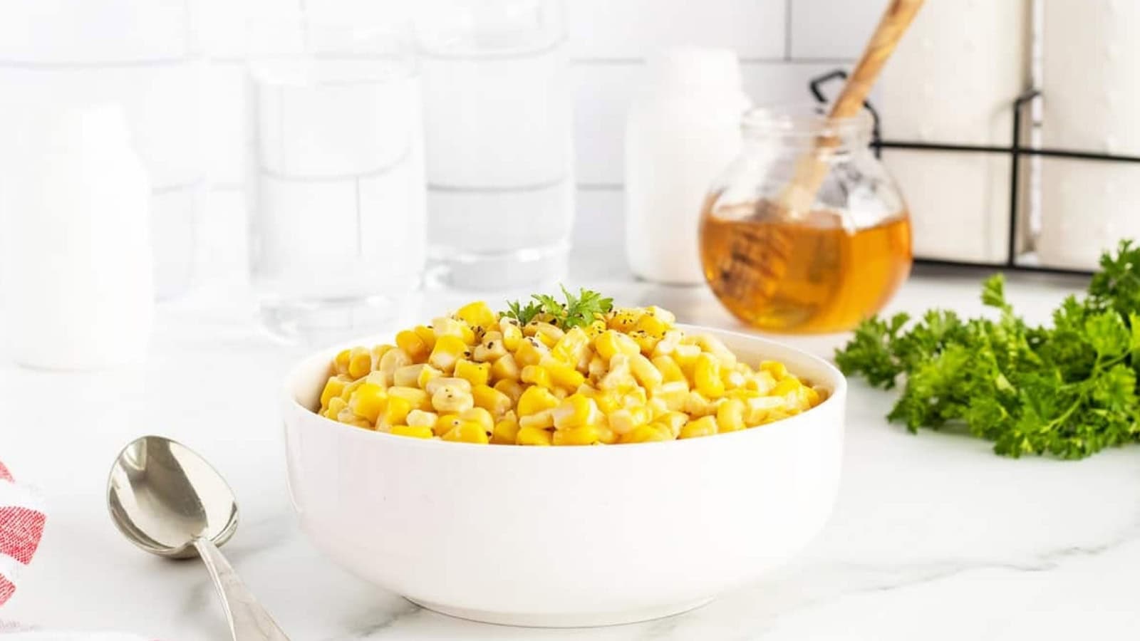 A white bowl of seasoned corn kernels garnished with parsley on a white counter, with honey and parsley nearby.