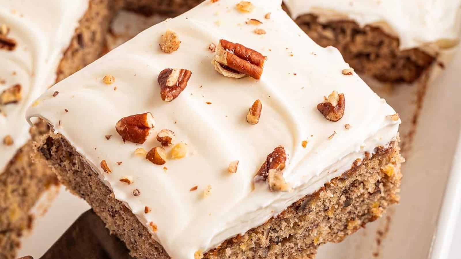 A slice of spiced cake topped with white frosting and chopped pecans, with more cake in the background.