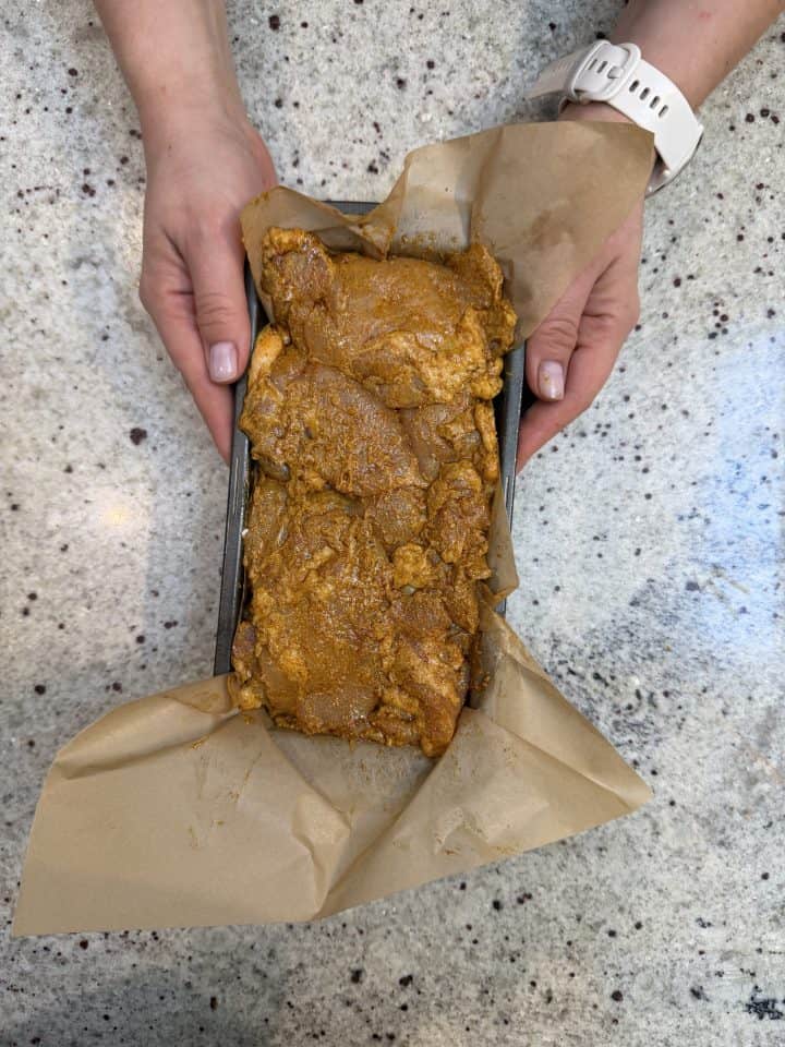 Hands holding a loaf pan lined with brown parchment paper and filled with marinated Loaf Pan Chicken Shawarma pieces.