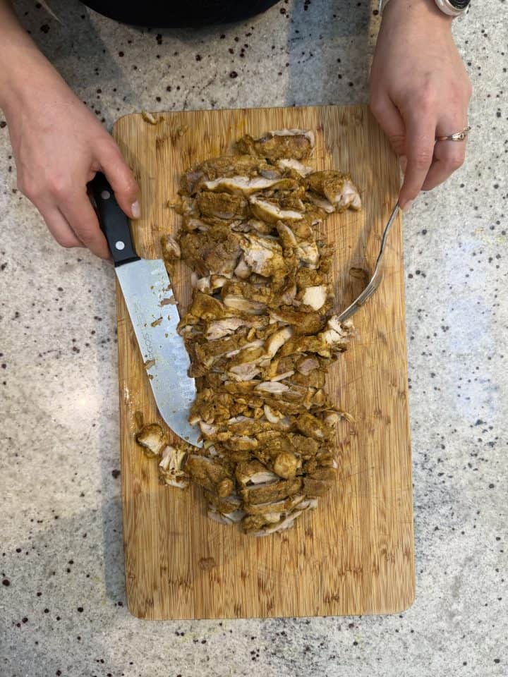Hands chopping seasoned cooked chicken—prepared as a flavorful Loaf Pan Chicken Shawarma—on a wooden cutting board with a knife and fork on a counter.