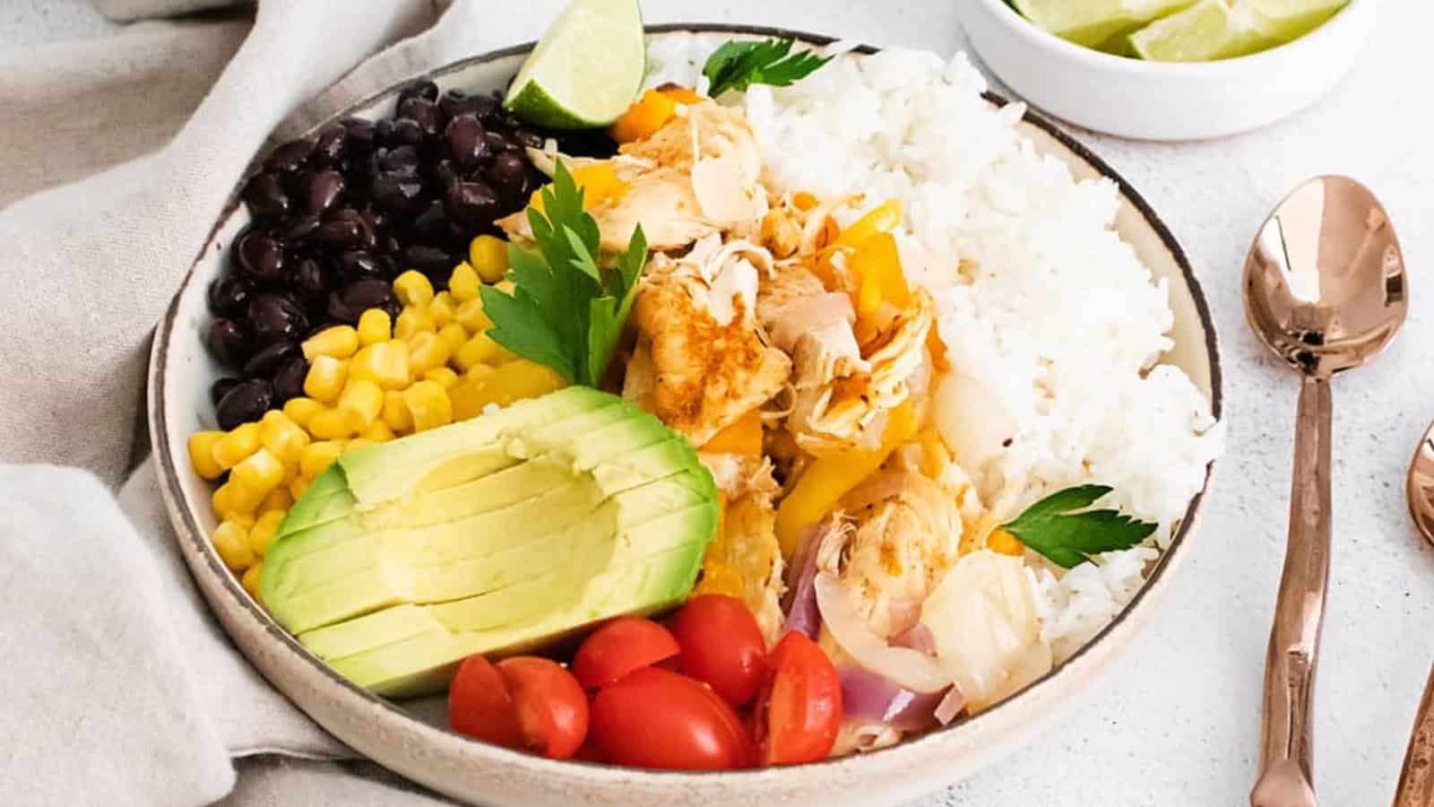A bowl with rice, shredded chicken, black beans, corn, avocado, cherry tomatoes, and a lime wedge.