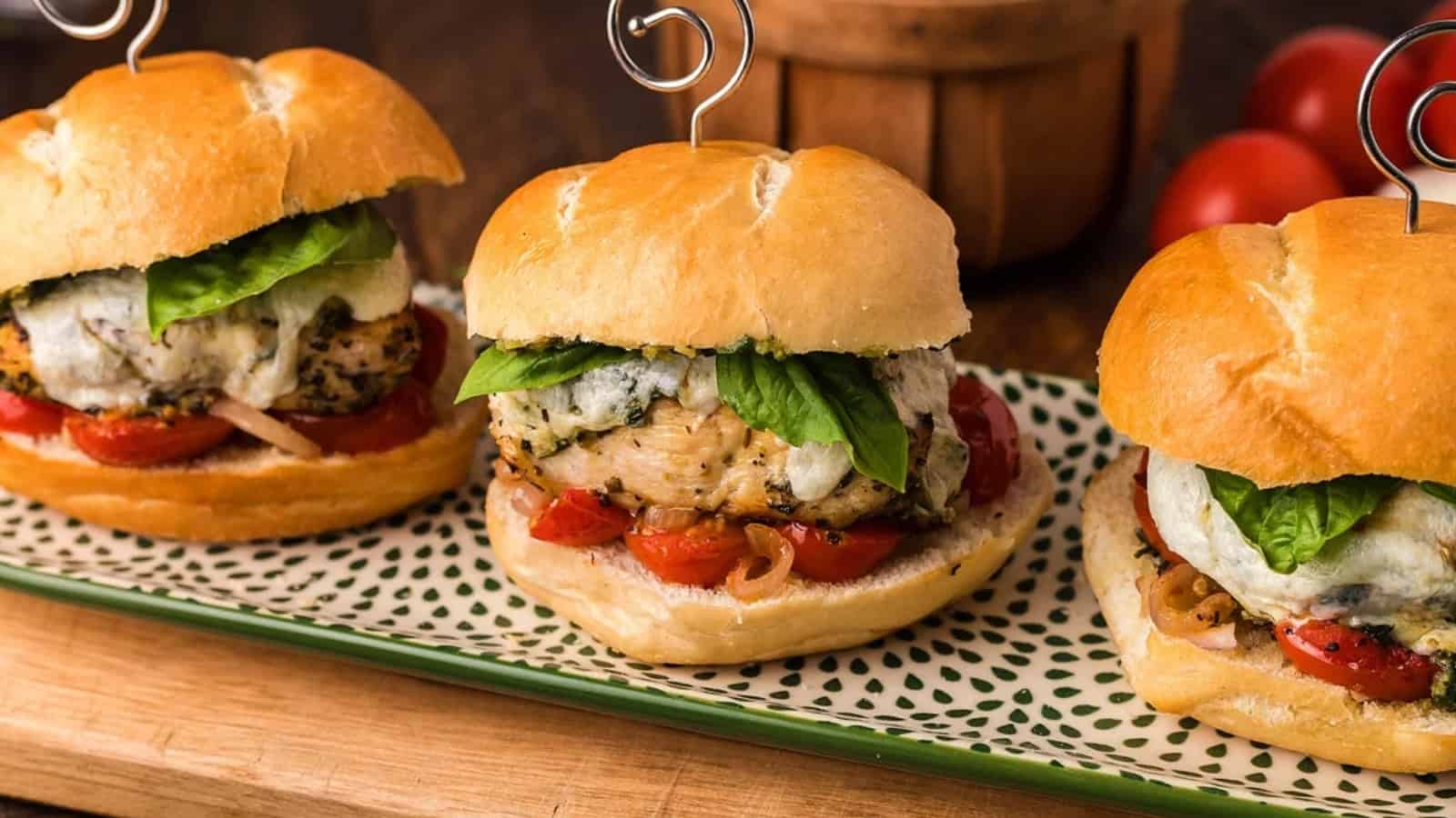 Three chicken burgers on a patterned plate, topped with cheese, fresh basil, and tomatoes.