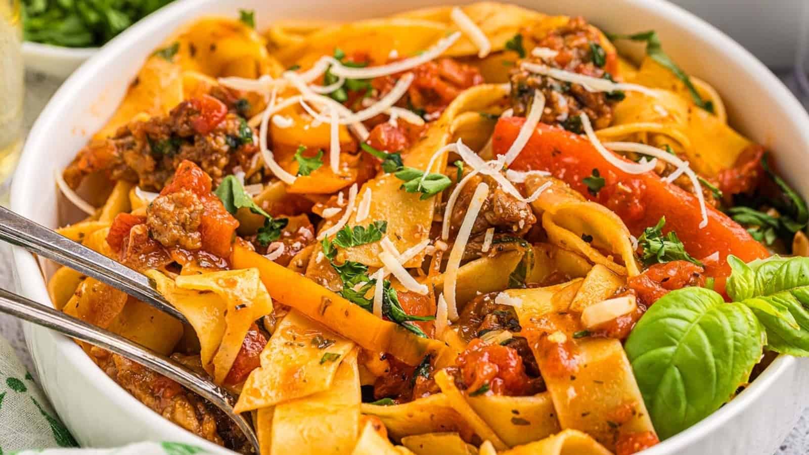 Wide pasta noodles with meat sauce, tomatoes, herbs, and grated cheese in a white bowl, garnished with basil.