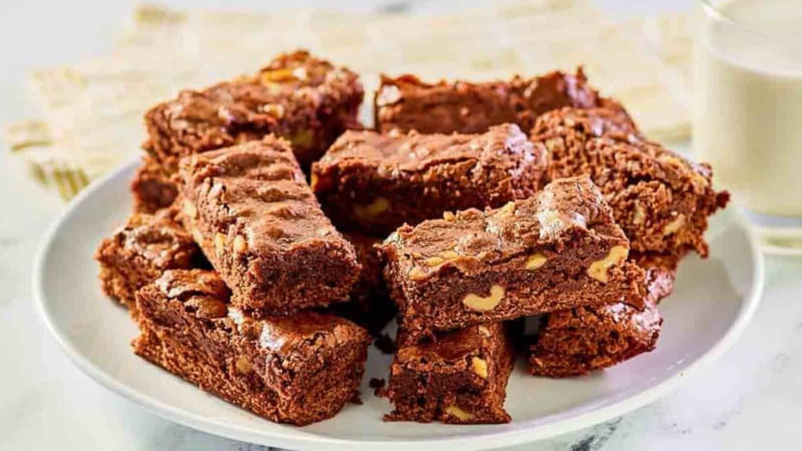 A plate of chocolate brownies with nuts, stacked beside a glass of milk on a white surface.