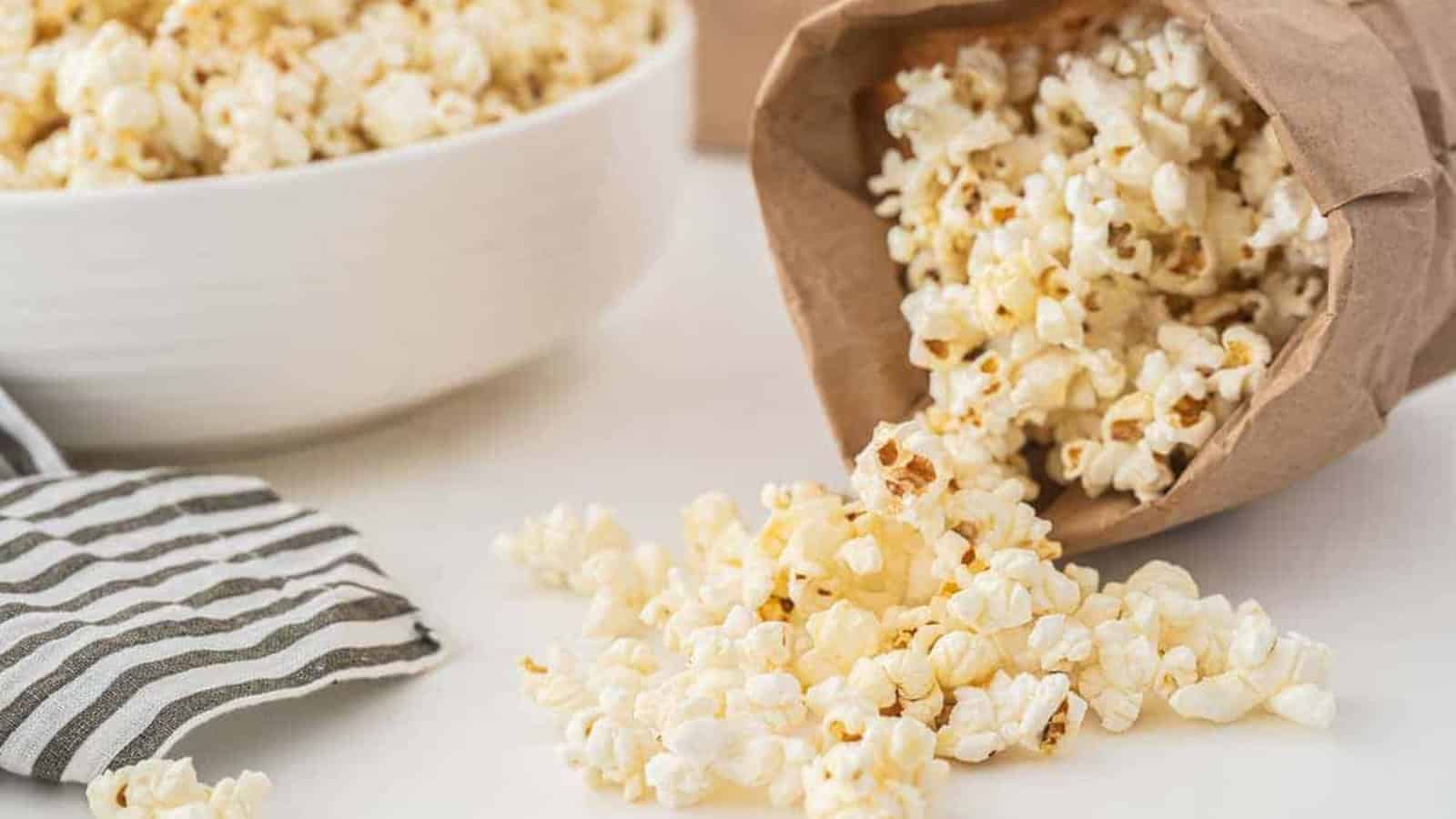 Popcorn spilling from a brown paper bag onto a table, with a bowl of popcorn and striped napkin nearby.