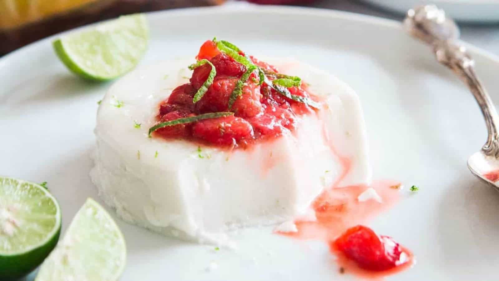 A panna cotta dessert topped with strawberry sauce and lime zest, served with lime wedges on the side.