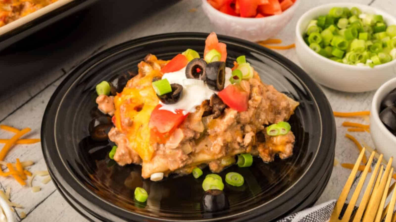 A serving of taco casserole topped with sour cream, tomatoes, olives, and green onions on a black plate.