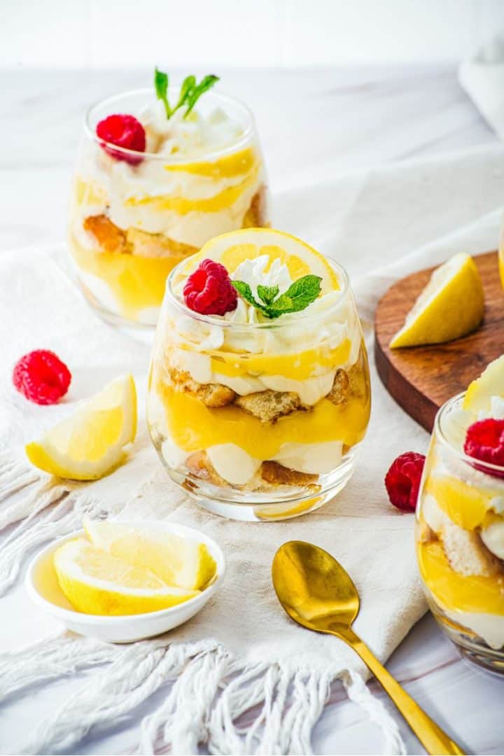 Lemon Cheesecake Trifle Cups in glasses, topped with lemon slices, raspberries, and mint, with gold spoons nearby.