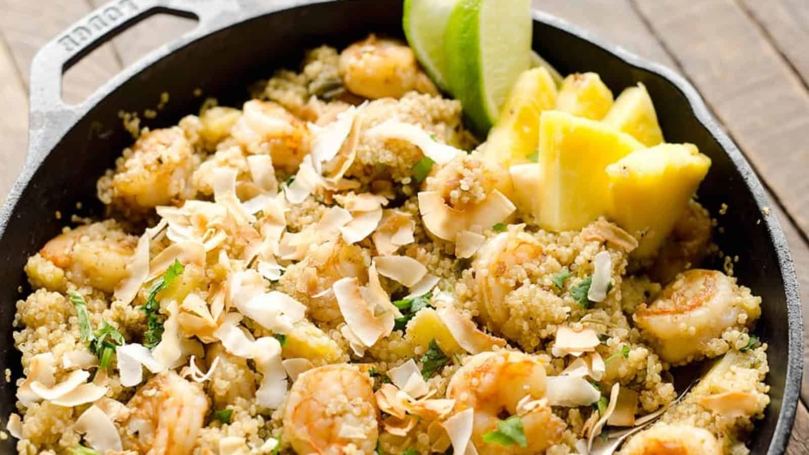 Shrimp, quinoa, toasted coconut, and pineapple chunks in a skillet, garnished with lime wedges.