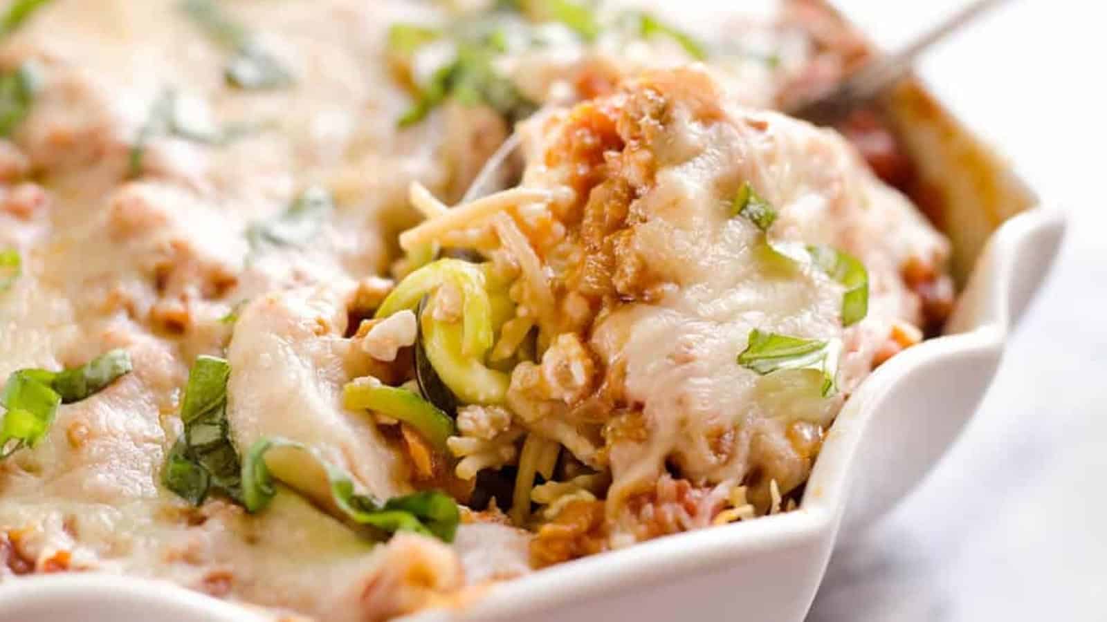 A fork lifts cheesy spaghetti bake topped with melted cheese and basil from a white dish.