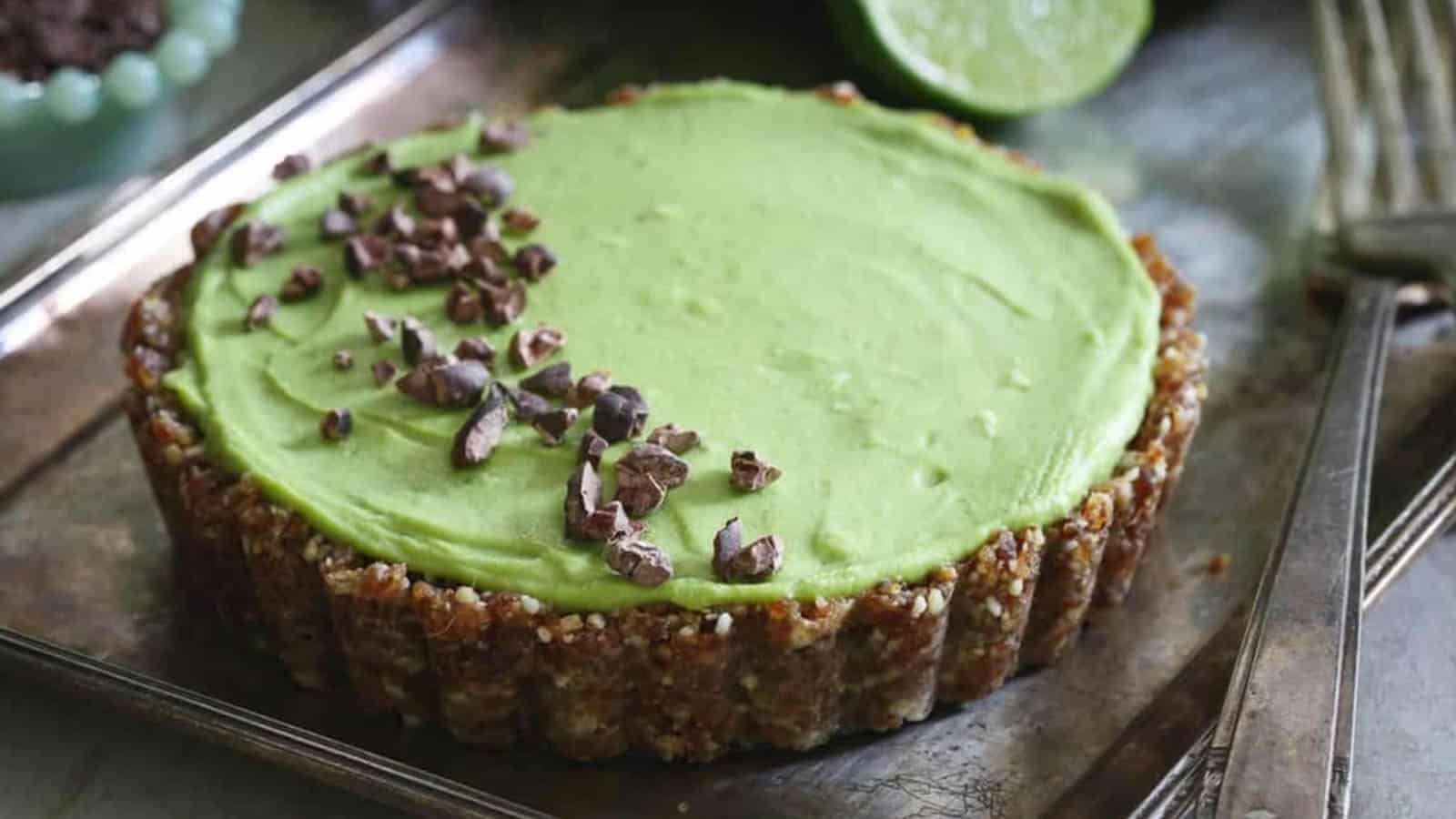 Green tart topped with cacao nibs on a metal tray, with a fork and sliced lime in the background.