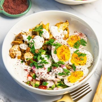 A bowl with soft-boiled eggs, grilled chicken, salad, yogurt sauce, fresh herbs, and spices.