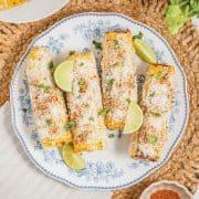 Four delicious Mexican street corn elotes on a plate, topped with cheese, cilantro, and lime wedges.