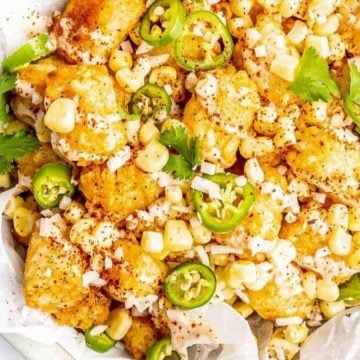 Tater tots topped with corn, jalapeños, cheese, cilantro, and a side of dipping sauce on a plate.