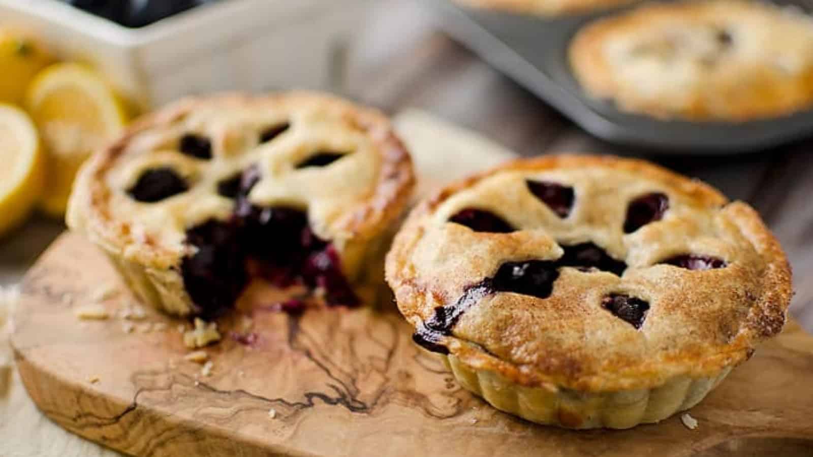 Two mini blueberry pies on a wooden board, one with a slice removed, showing the filling.