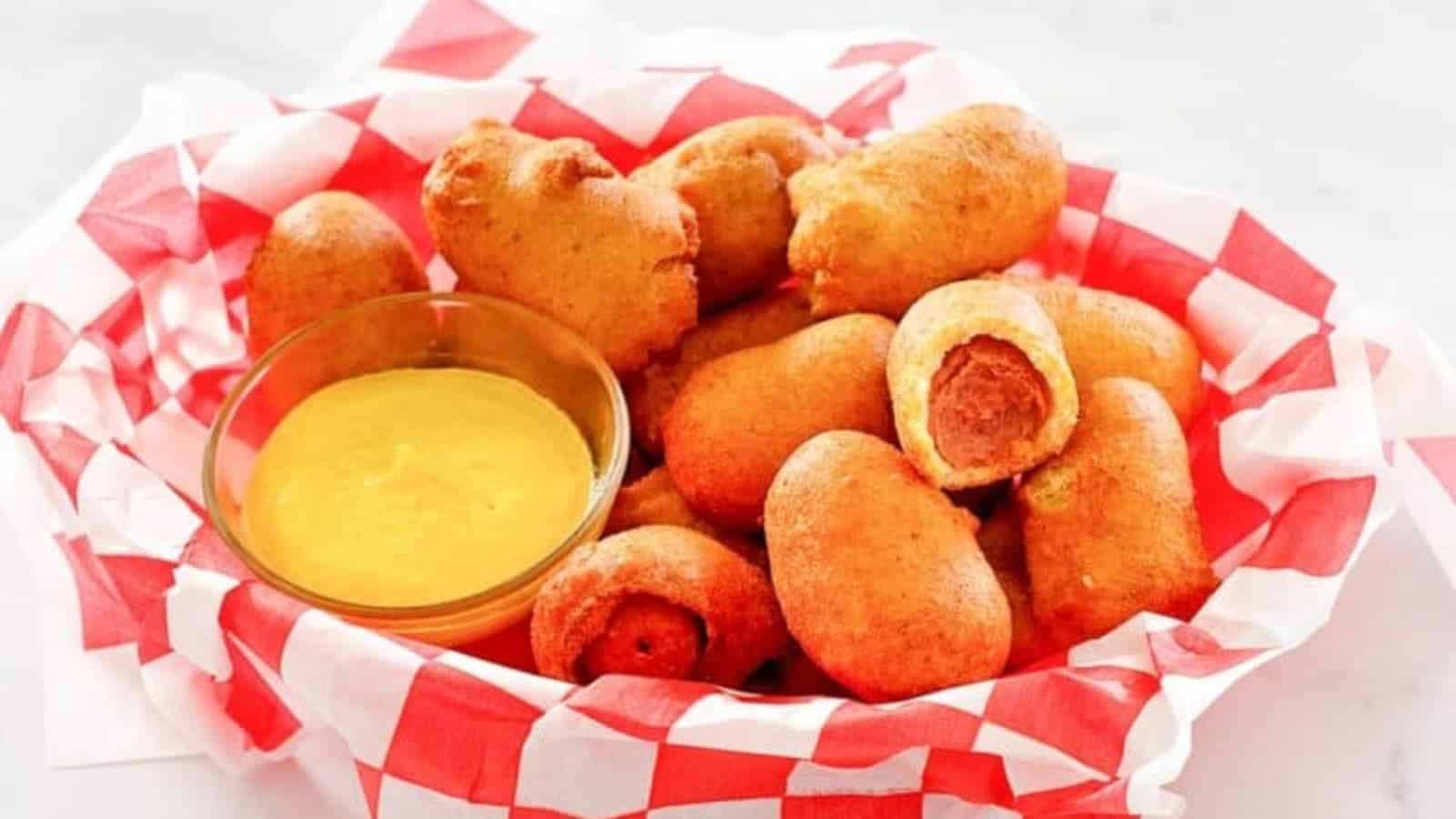 Mini corn dogs in a red and white basket with a bowl of yellow mustard dipping sauce.