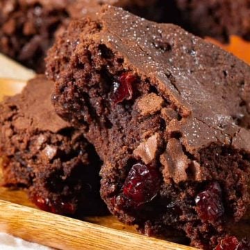 Two chocolate brownies with visible cranberries on a wooden tray, surrounded by autumn leaves.