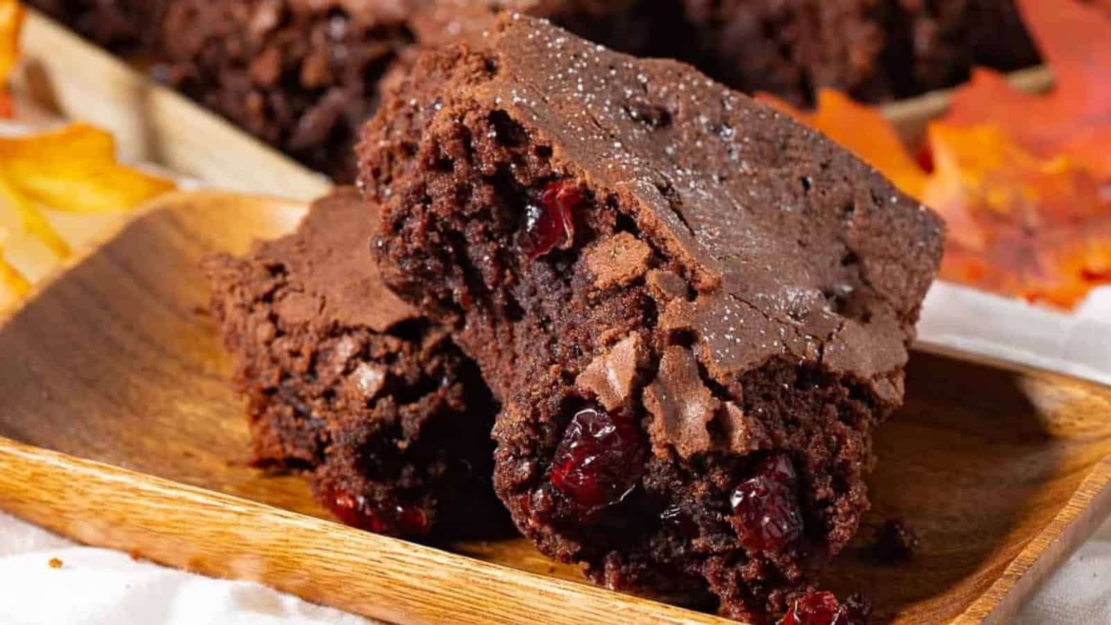Two chocolate brownies with visible cranberries on a wooden tray, surrounded by autumn leaves.