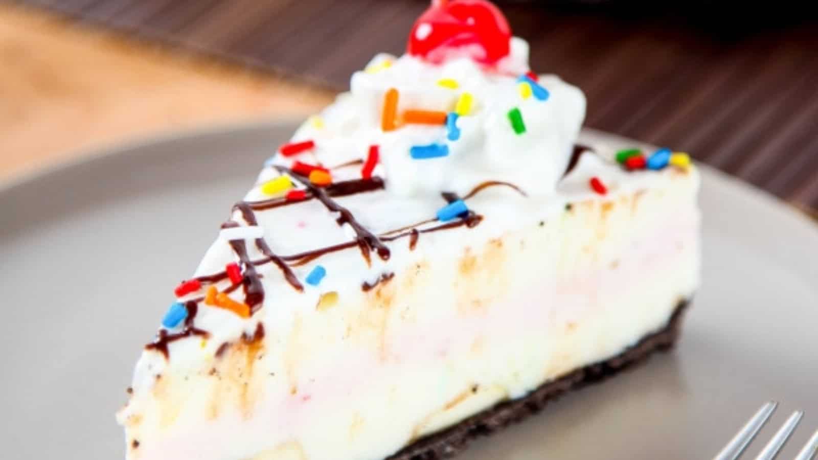 A slice of ice cream cake with whipped cream, sprinkles, chocolate drizzle, and a cherry on top.