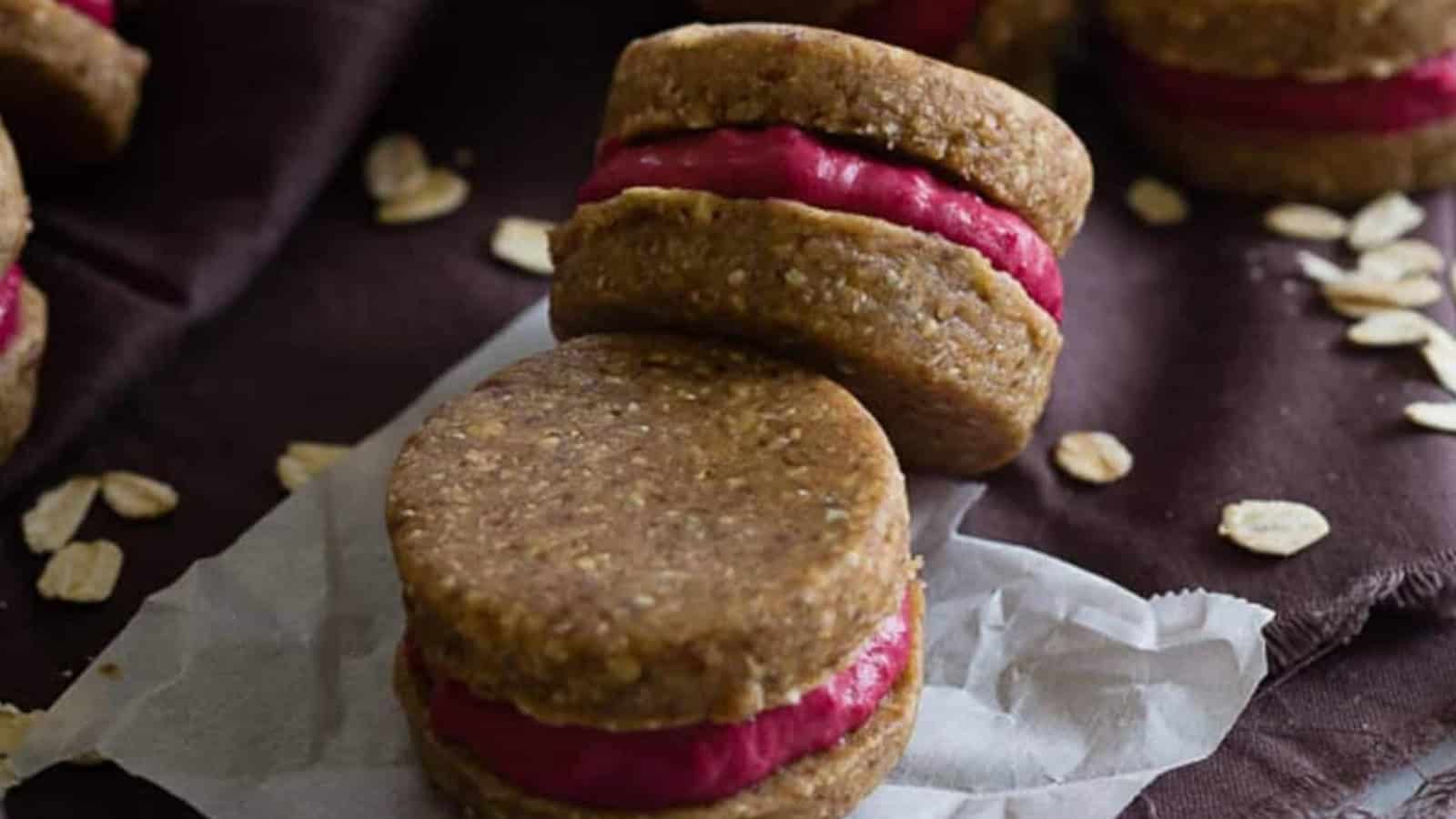 Two oatmeal sandwich cookies with bright pink filling, set on parchment paper with oats scattered nearby.