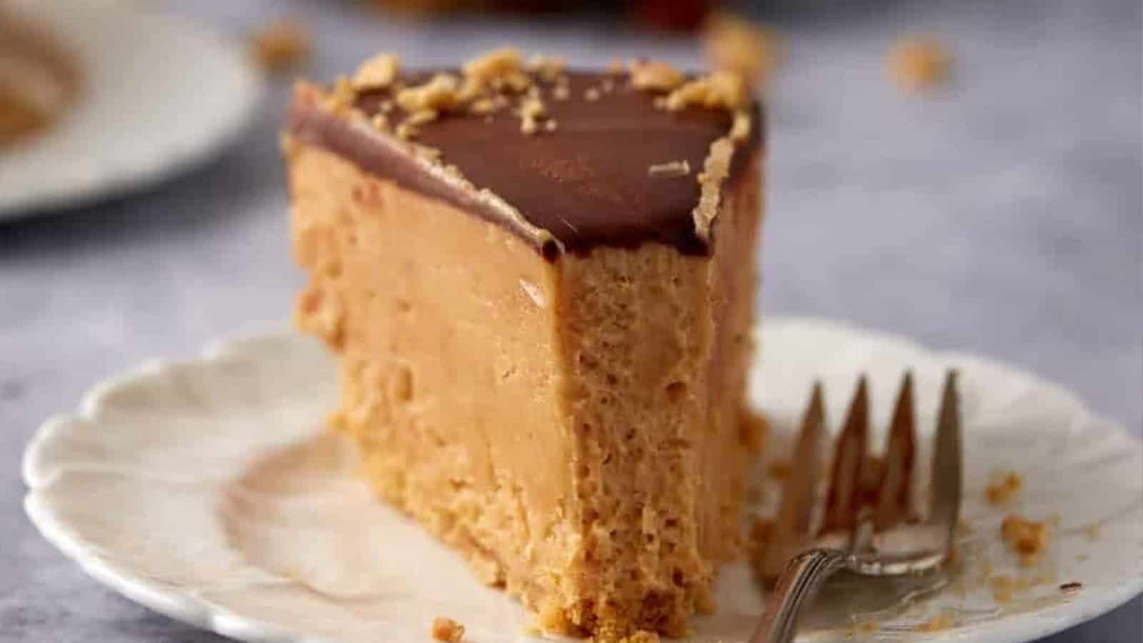 A slice of peanut butter cheesecake with chocolate topping on a white plate and a fork beside it.