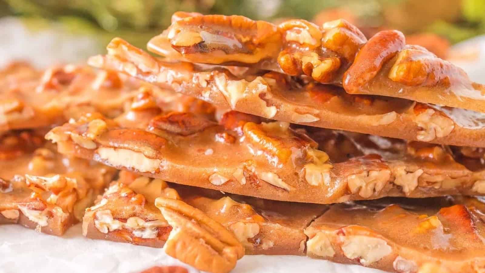 Stack of golden brown pecan brittle pieces with visible whole and chopped pecans on a white surface.