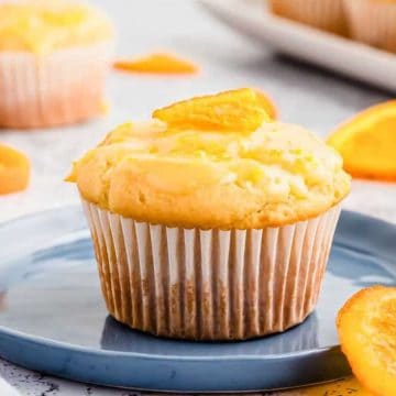 Orange muffin on a blue plate with orange slices, more muffins and orange pieces blurred in the background.