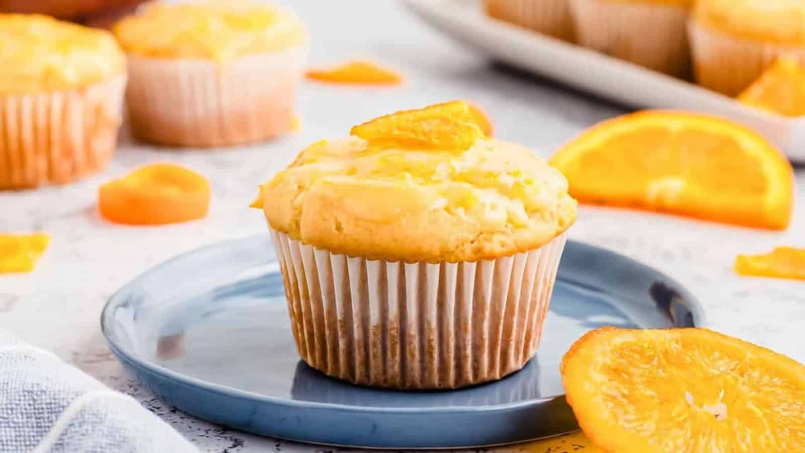 Orange muffin on a blue plate with orange slices, more muffins and orange pieces blurred in the background.